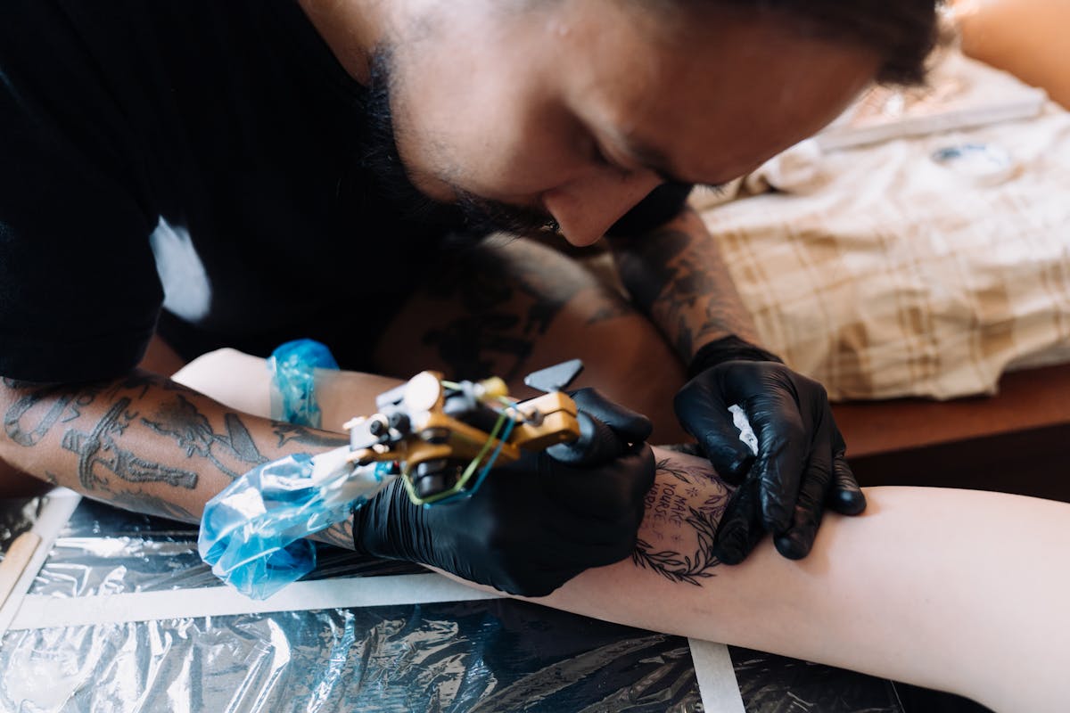 Close-up of a tattoo artist working on a forearm piece, ink and gloves visible.