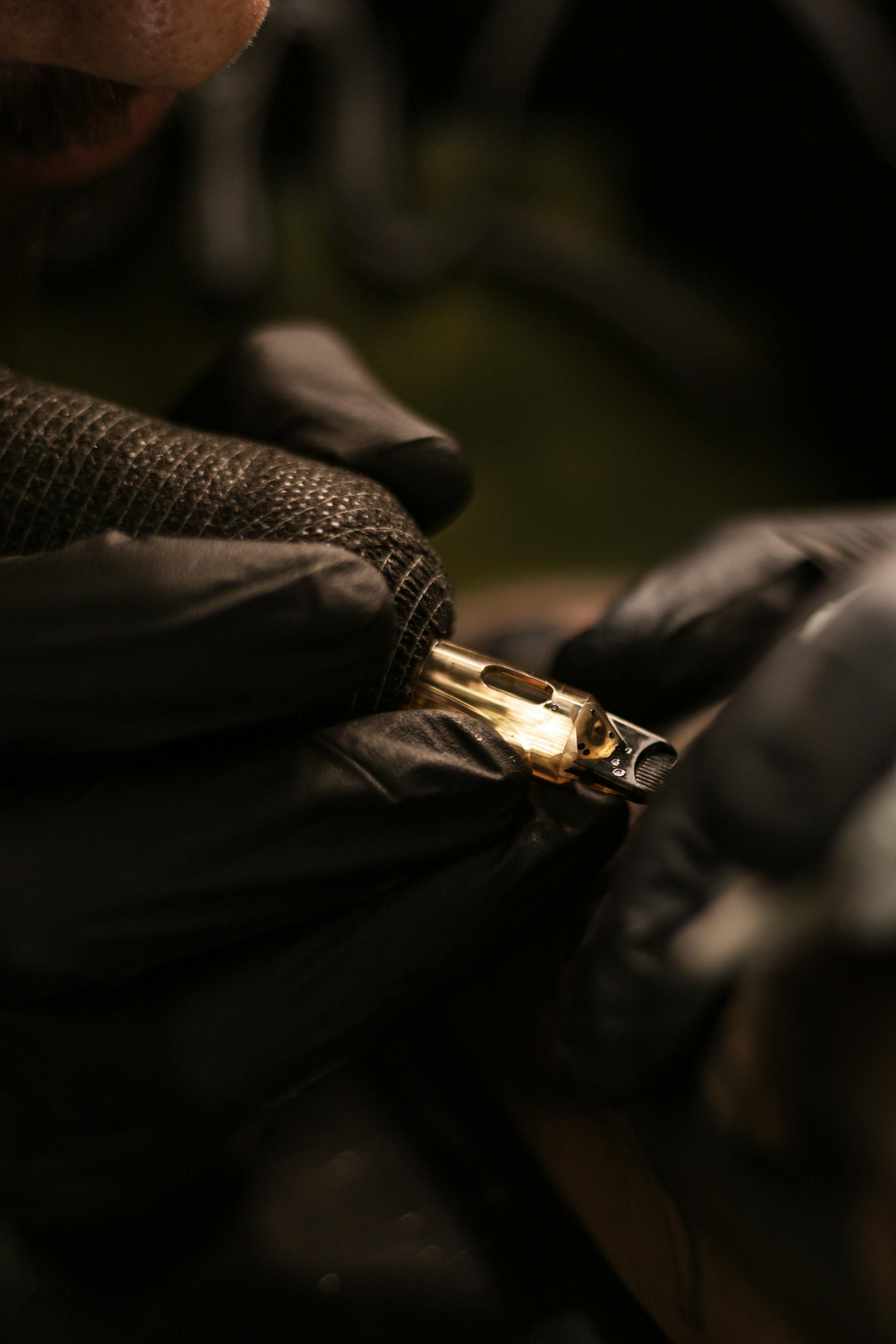 Close-up of black-gloved hands holding a brass tattoo machine in warm low light — focused, deliberate work.